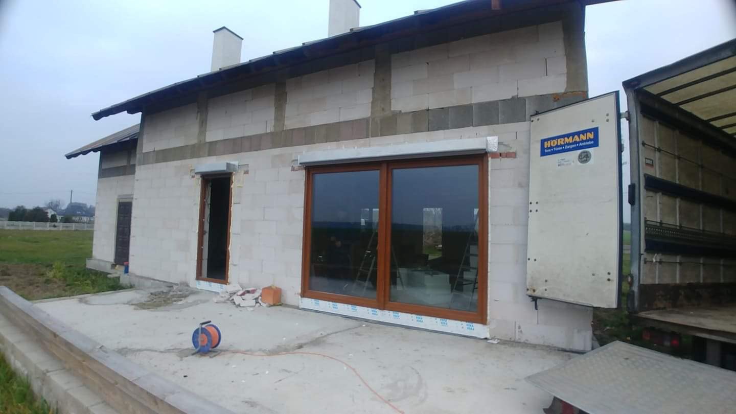 Surowy stan budynku w trakcie montażu stolarki okiennej, widoczne okno dwuskrzydłowe z brązowymi ramami, częściowo opuszczona roleta zewnętrzna oraz otwarte drzwi wejściowe, na zewnątrz zaparkowana...