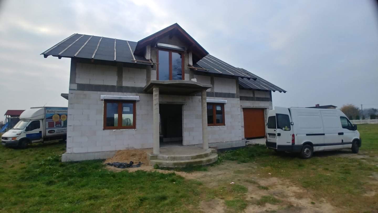 Dom w stanie surowym zamkniętym w Ostrowie Wielkopolskim z zamontowanymi oknami z brązowymi ramami i częściowo zamontowanymi roletami zewnętrznymi, widoczna brama garażowa i dwa samochody dostawcze.