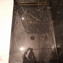 Wiktor Kharchenko usługi budowlane - Wykończona strefa prysznicowa z czarnych płytek imitujących marmur z złotymi żyłami, z odpływem liniowym w kolorze złotym i złotym deszczownikiem, widoczne odbicie osoby robiącej zdjęcie i klapki...