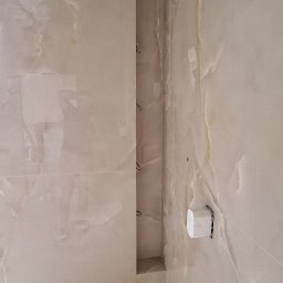 Wiktor Kharchenko usługi budowlane - Ściana wyłożona beżowymi płytkami imitującymi marmur z wnęką, widoczny fragment sufitu z płytami gipsowo-kartonowymi oraz uchwyt na papier toaletowy.