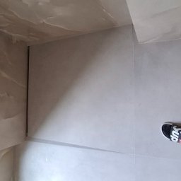 Wiktor Kharchenko usługi budowlane - Wykończona łazienka z beżowymi płytkami imitującymi marmur na ścianach i podłodze, widoczne stopy w czarnych skarpetach i klapkach Supreme.