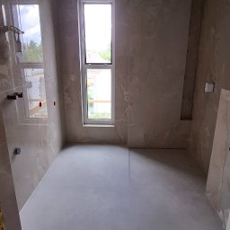 Wiktor Kharchenko usługi budowlane - Wnętrze łazienki w trakcie prac wykończeniowych, widoczne płytki imitujące marmur na ścianach, sufit z płyt gipsowo-kartonowych, okno z widokiem na zieleń oraz przygotowana podłoga.