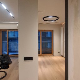 Wiktor Kharchenko usługi budowlane - Jasne panele podłogowe w nowoczesnym wnętrzu z oknami i minimalistycznym oświetleniem sufitowym. Widoczny fragment kolumny z włącznikami.