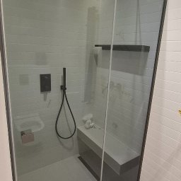 Wiktor Kharchenko usługi budowlane - Nowoczesna kabina prysznicowa z grafitowymi elementami, szklanymi drzwiami i subtelnym oświetleniem LED. Widoczna półka i siedzisko wewnątrz kabiny.