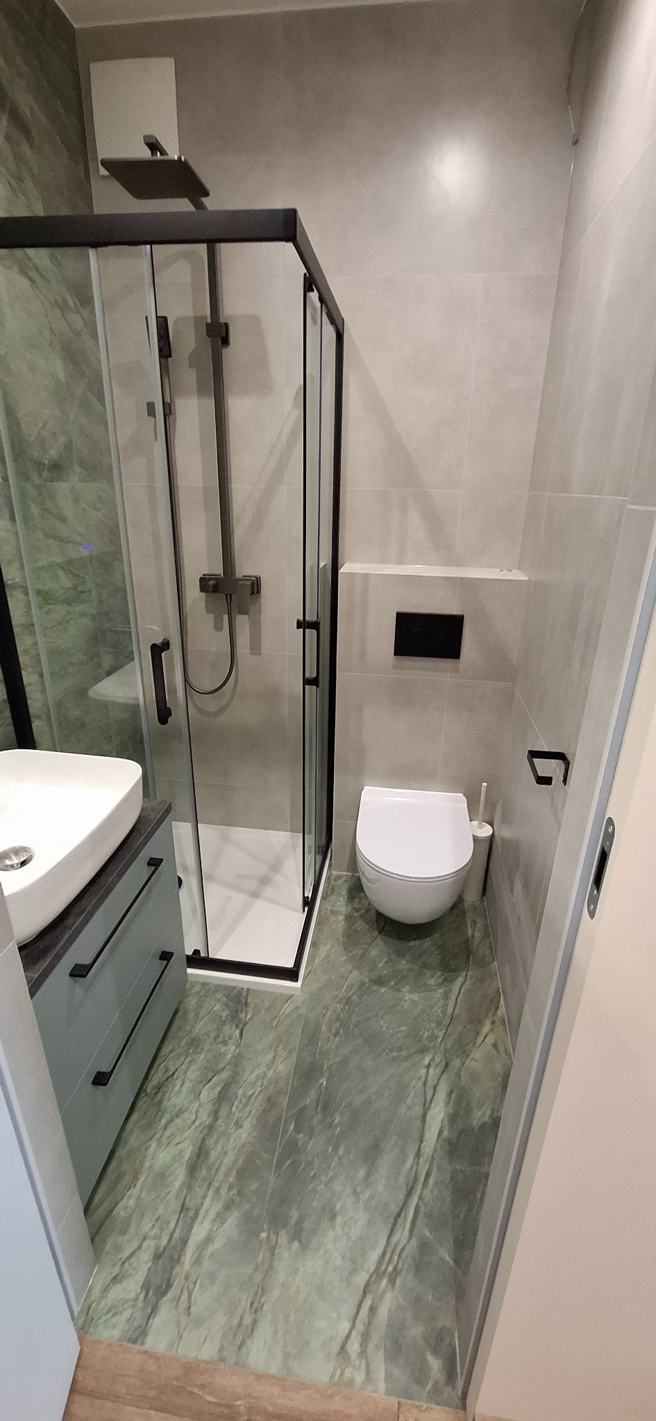 Nowoczesna łazienka z kabiną prysznicową typu walk-in z czarnymi profilami, umywalką nablatową na szafce z szufladami w kolorze gołębim, podwieszaną toaletą i podłogą z płytek imitujących zielony...