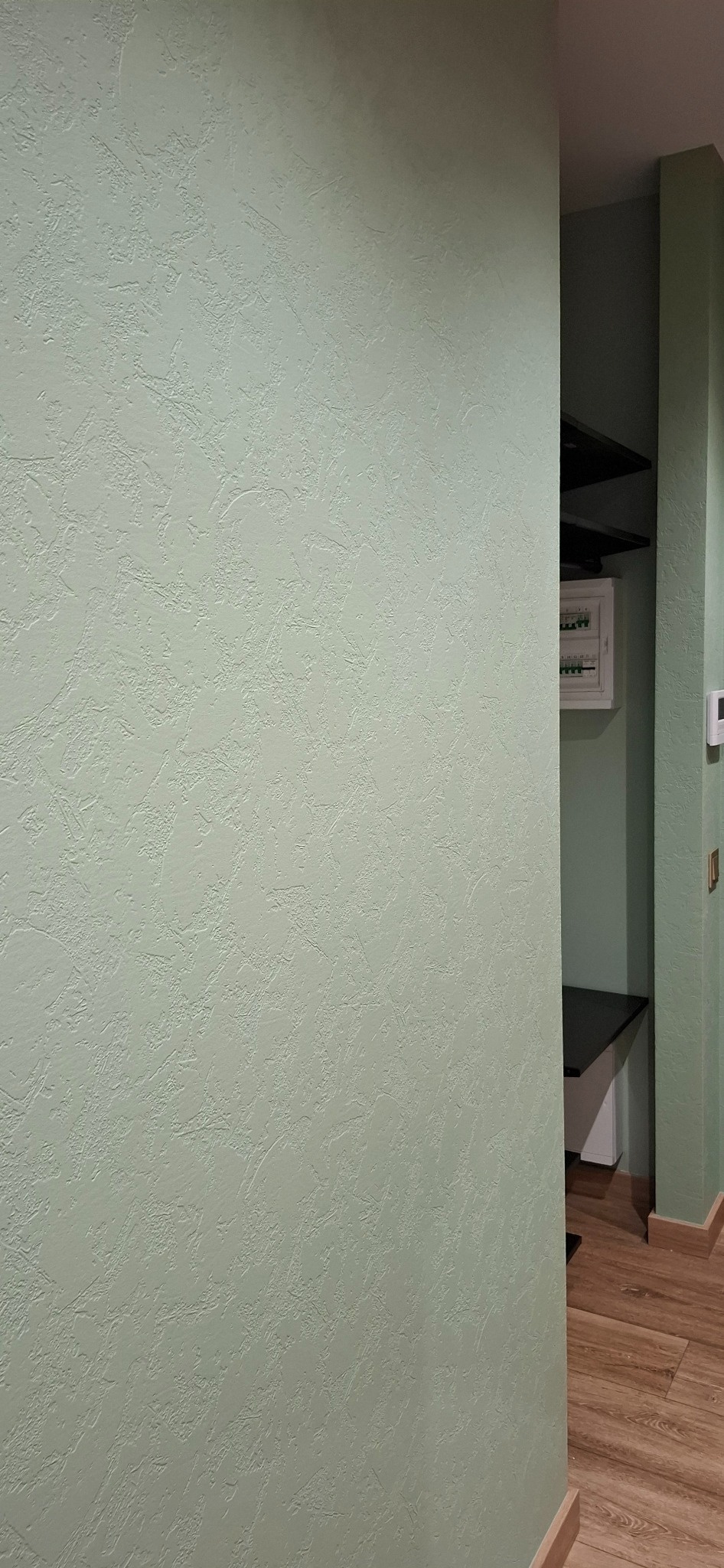 Jasnozielona ściana z dekoracyjną fakturą, widoczny fragment pomieszczenia z szafkami i skrzynką elektryczną w tle.