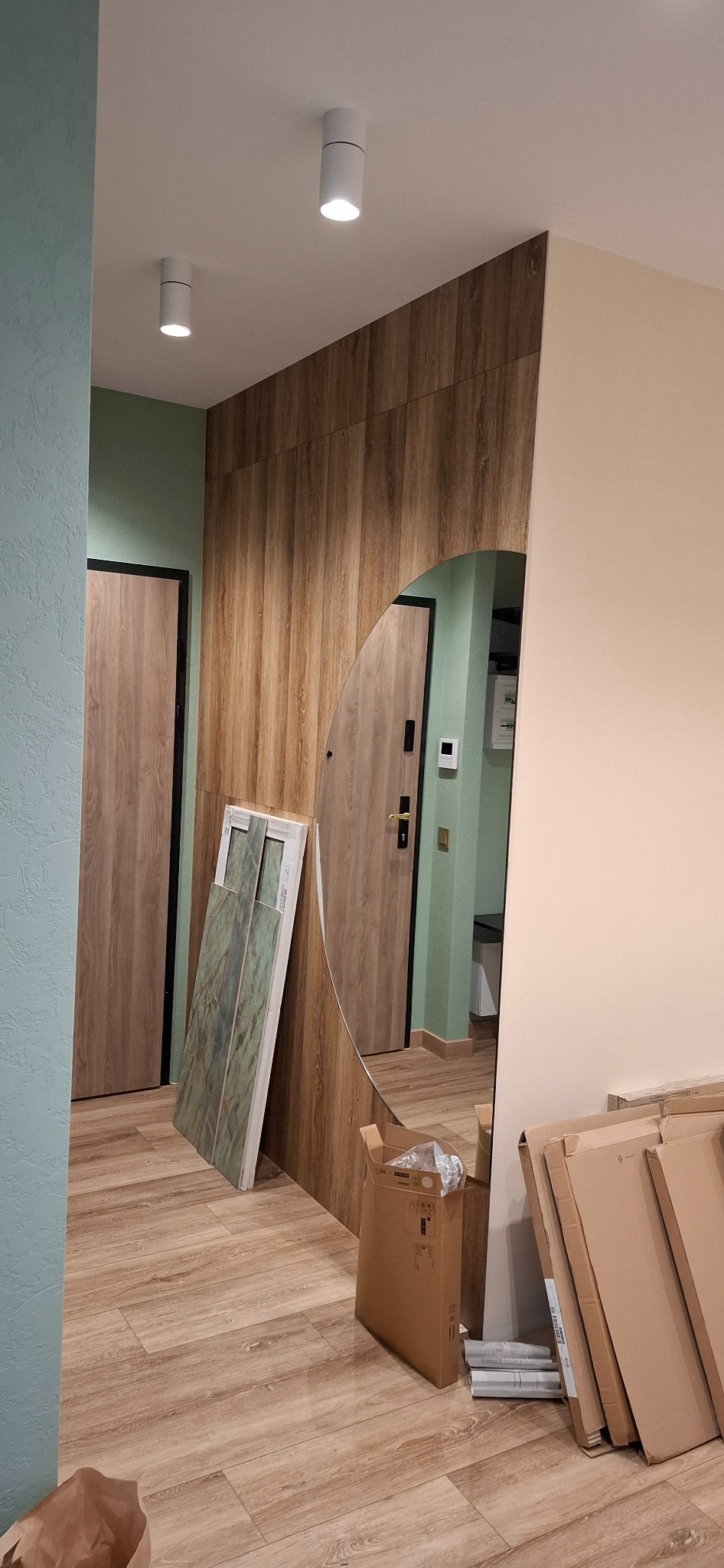 Korytarz w trakcie prac wykończeniowych: położona podłoga z paneli imitujących drewno, zielone płytki ceramiczne oparte o ścianę, okrągłe lustro, kartony po panelach i drzwi z czarną ościeżnicą.