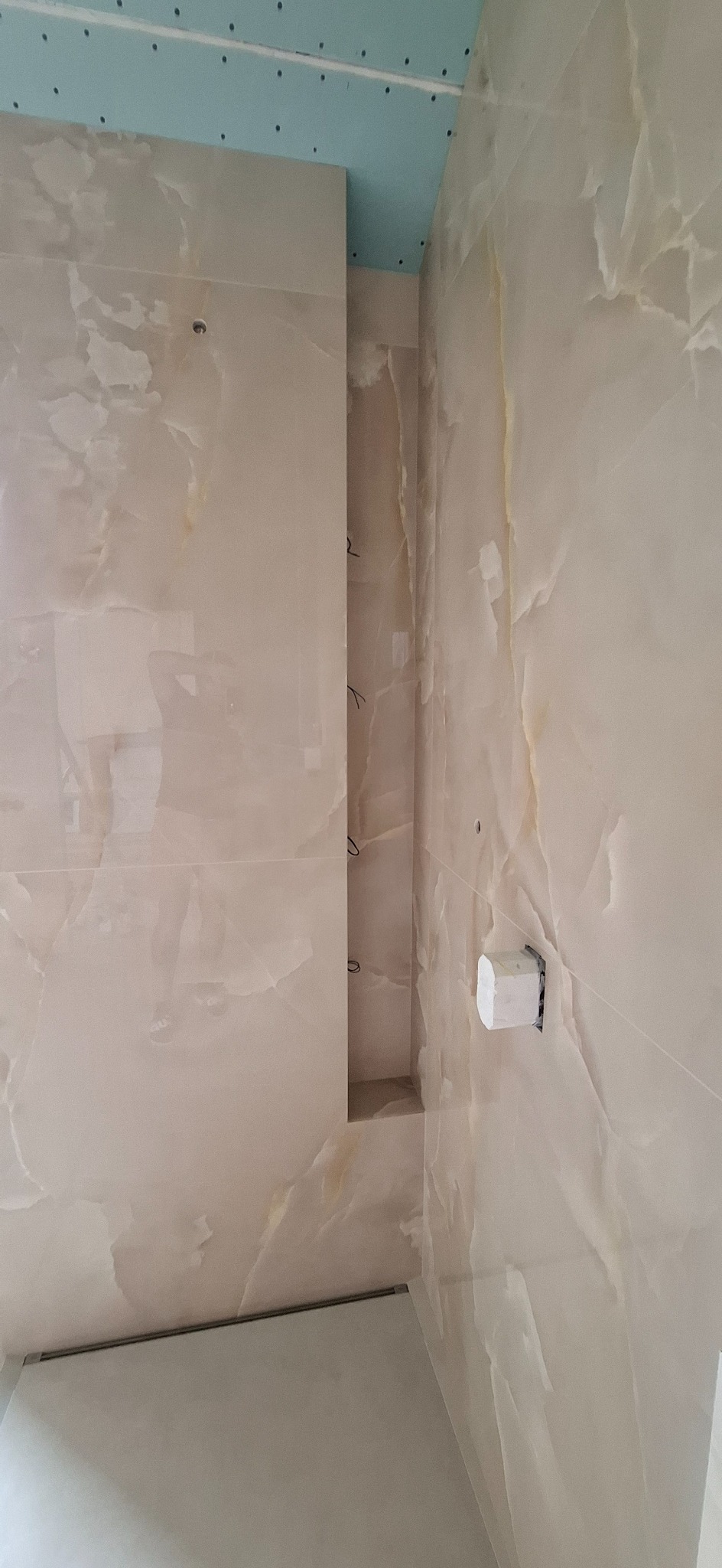 Ściana wyłożona beżowymi płytkami imitującymi marmur z wnęką, widoczny fragment sufitu z płytami gipsowo-kartonowymi oraz uchwyt na papier toaletowy.