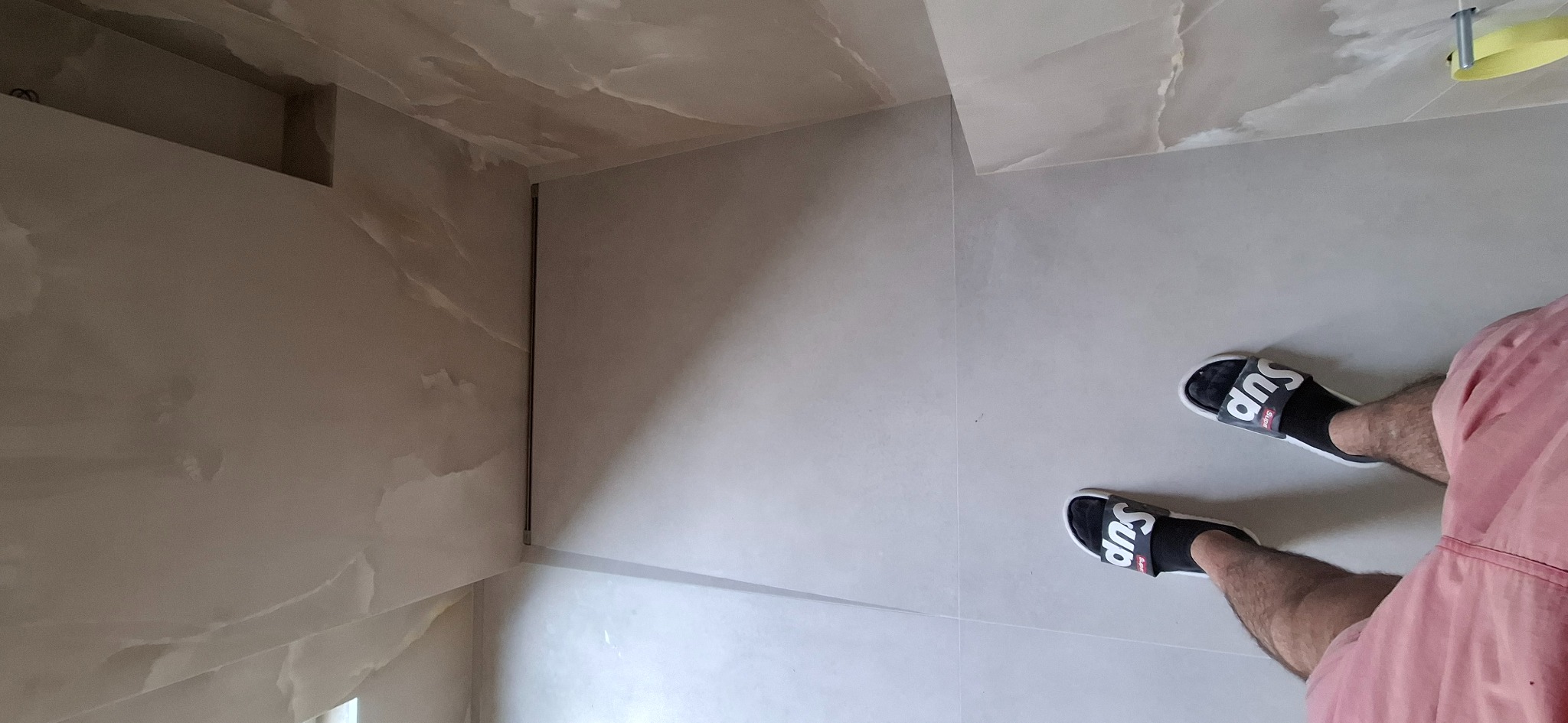 Wykończona łazienka z beżowymi płytkami imitującymi marmur na ścianach i podłodze, widoczne stopy w czarnych skarpetach i klapkach Supreme.