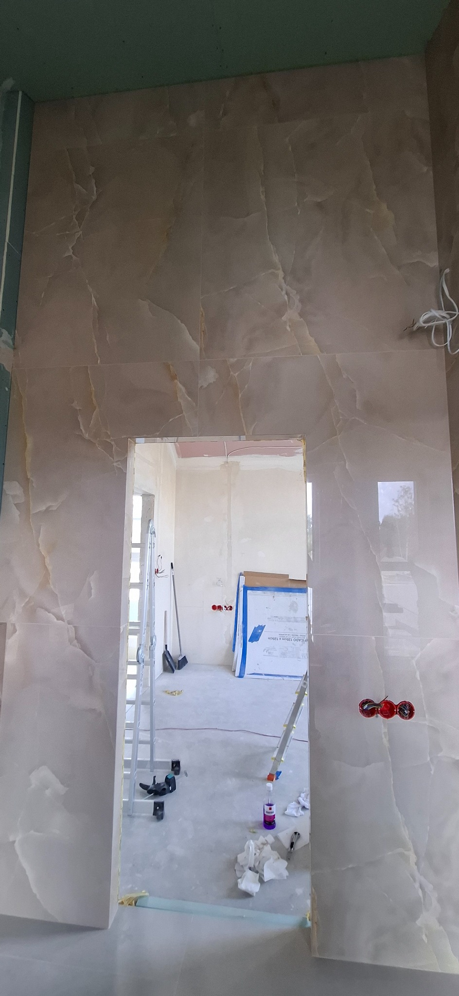 Ściana w trakcie wykańczania płytkami imitującymi marmur, widoczne narzędzia, drabina i przygotowane materiały budowlane w pomieszczeniu obok.
