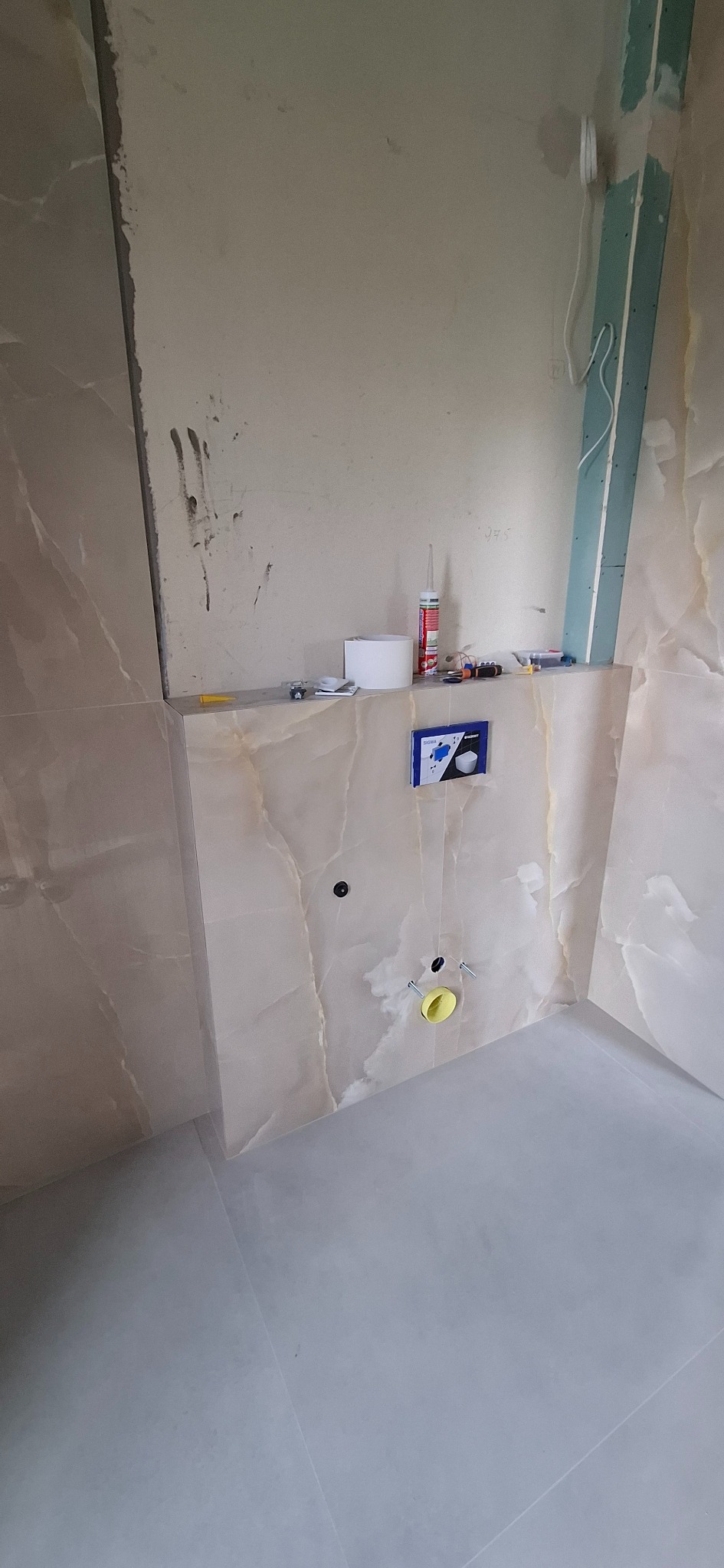 Wnętrze łazienki w trakcie prac wykończeniowych; widoczne płytki imitujące marmur na ścianach, przygotowane instalacje sanitarne oraz narzędzia i materiały budowlane na półce.