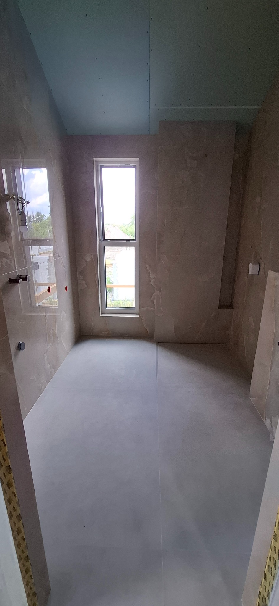 Wnętrze łazienki w trakcie prac wykończeniowych, widoczne płytki imitujące marmur na ścianach, sufit z płyt gipsowo-kartonowych, okno z widokiem na zieleń oraz przygotowana podłoga.