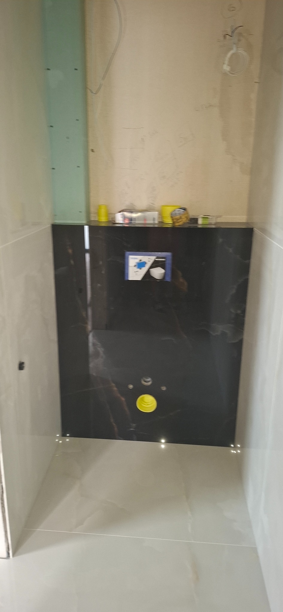 Stelaż podtynkowy z czarną płytą imitującą marmur, częściowo obudowany płytkami w odcieniach beżu, widoczne przyłącza i puszki z akcesoriami na górze.