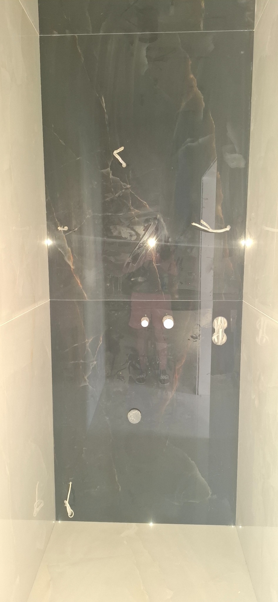 Ściana łazienki wyłożona płytkami imitującymi czarny marmur z widocznymi otworami na oświetlenie i wystającymi przewodami elektrycznymi przed montażem.