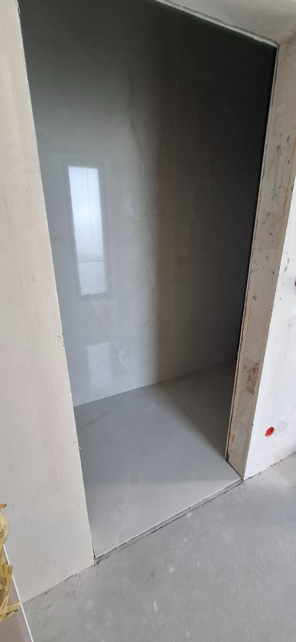 Wnętrze kabiny prysznicowej w trakcie wykańczania, widoczne białe płytki imitujące marmur na ścianach i podłodze, surowy beton na ścianach zewnętrznych.