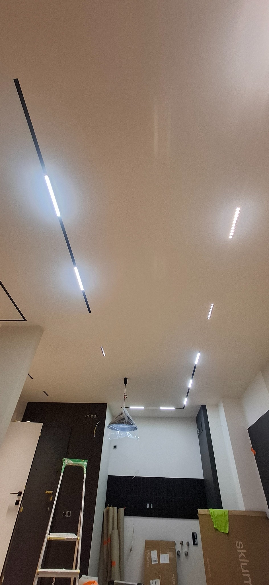 Nowoczesne oświetlenie sufitowe z liniowymi lampami LED w trakcie montażu. Widoczna drabina i materiały budowlane, co sugeruje etap prac wykończeniowych.