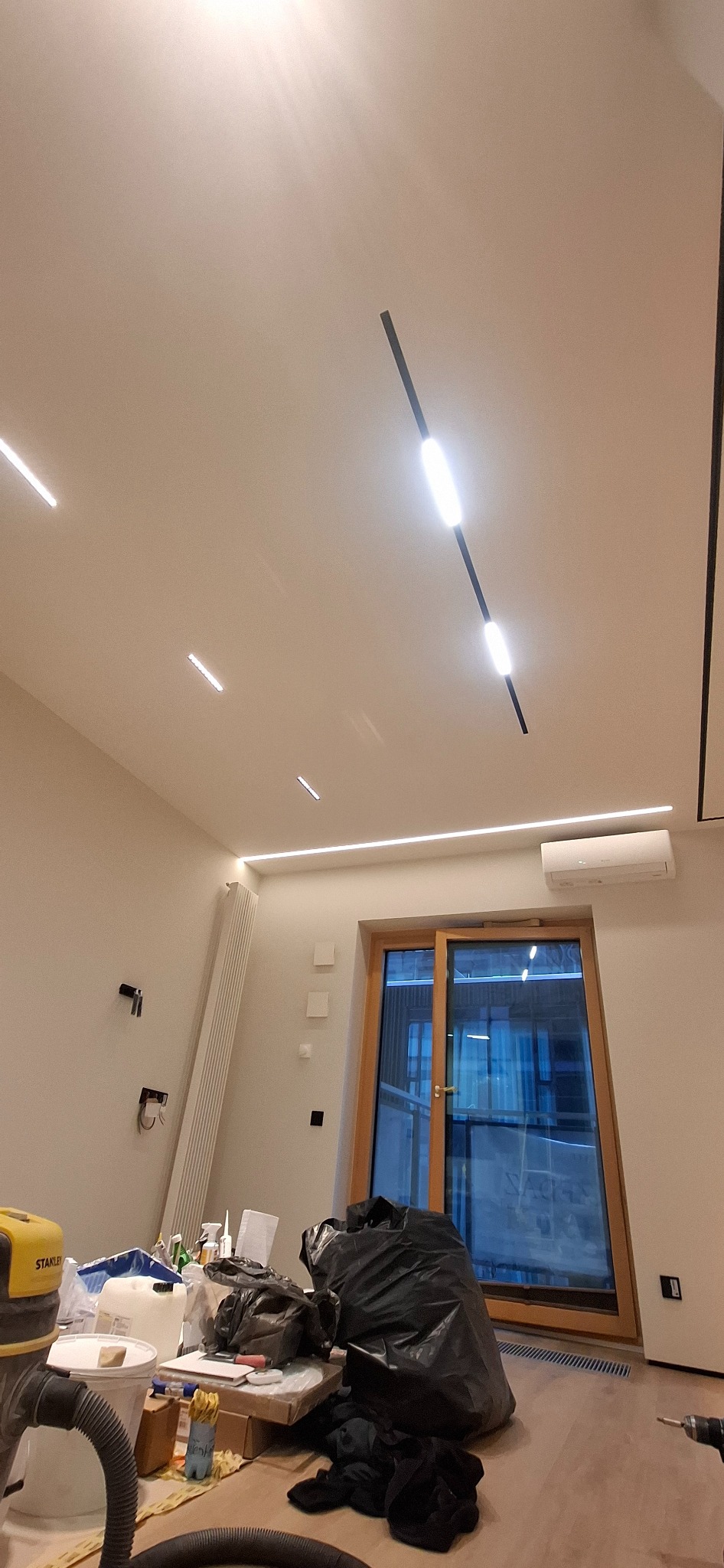 Wnętrze w trakcie prac: widoczne narzędzia, worki na śmieci, sufit z nowoczesnym oświetleniem liniowym i klimatyzacja. Częściowo widoczne okno balkonowe.