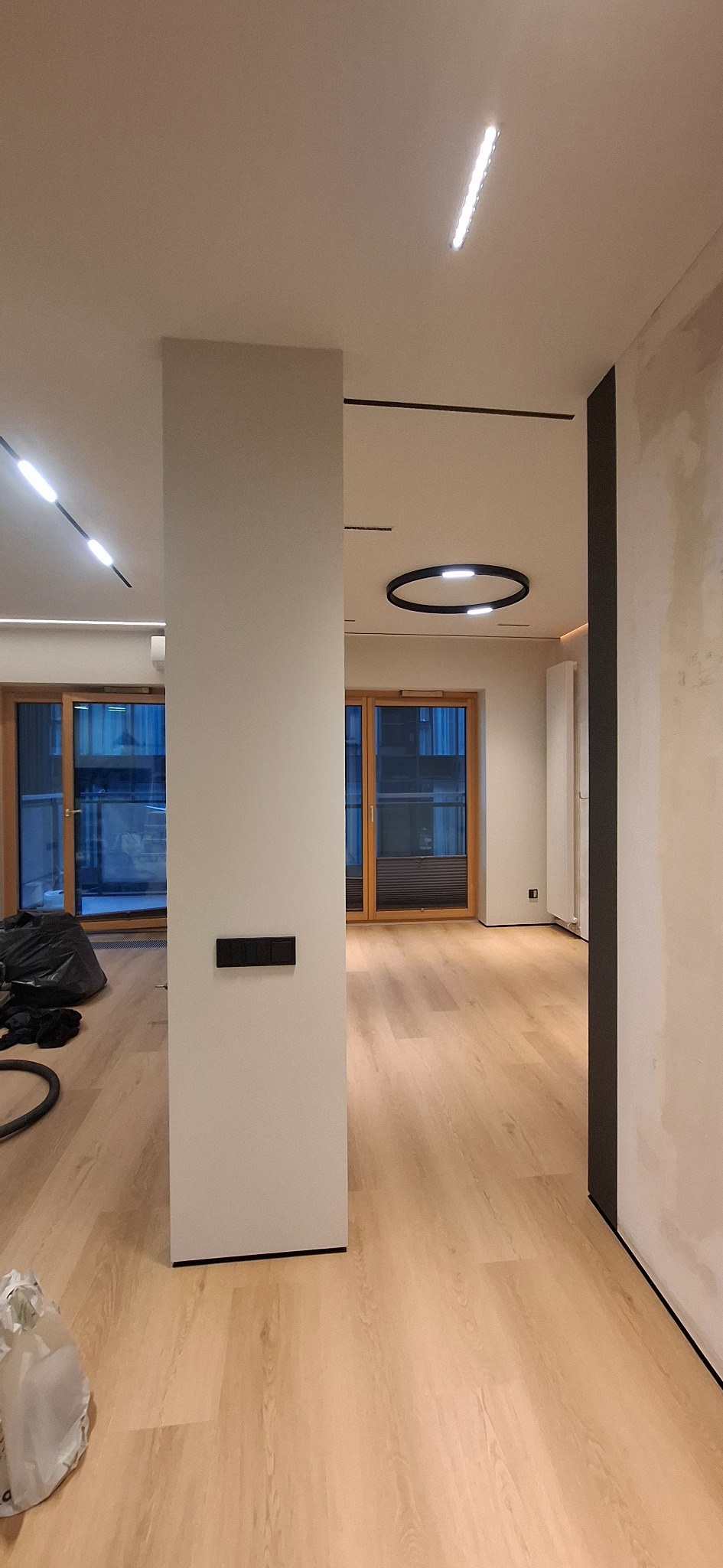 Jasne panele podłogowe w nowoczesnym wnętrzu z oknami i minimalistycznym oświetleniem sufitowym. Widoczny fragment kolumny z włącznikami.