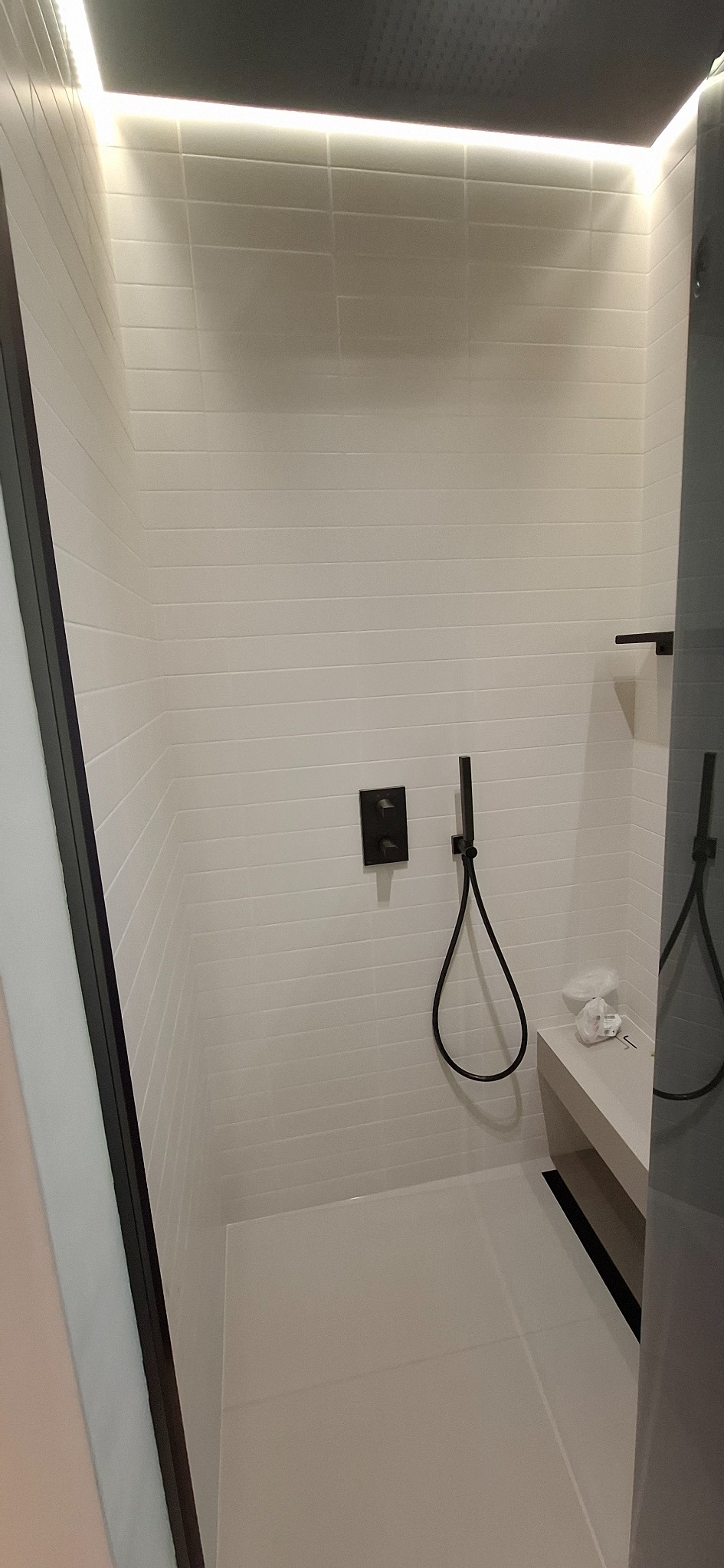 Nowoczesna kabina prysznicowa z białymi, poziomymi płytkami, czarną armaturą i oświetleniem LED. Minimalistyczny design z ławką i odpływem liniowym.