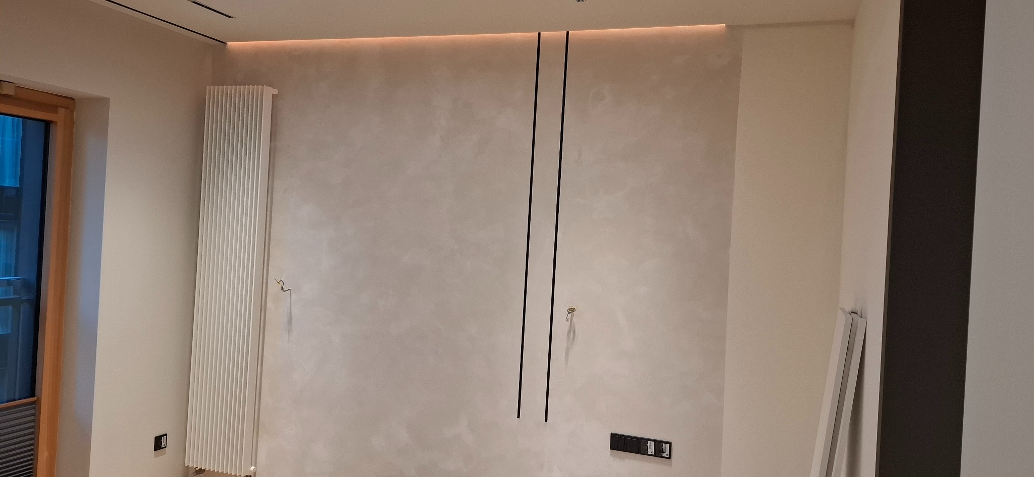 Wykończona ściana z dekoracyjnym tynkiem w odcieniach beżu, oświetleniem LED podsufitowym i pionowymi czarnymi listwami. Widoczny grzejnik i gniazdka elektryczne.