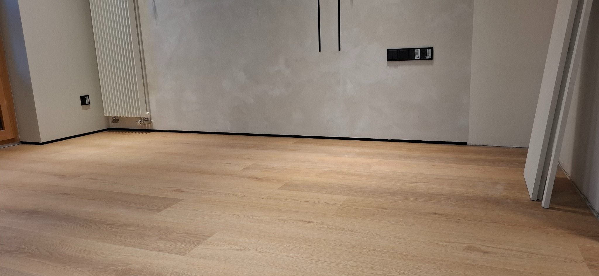 Jasne panele podłogowe ułożone w pokoju z grzejnikiem i szarą ścianą. Widoczne listwy przypodłogowe i fragment ościeżnicy.