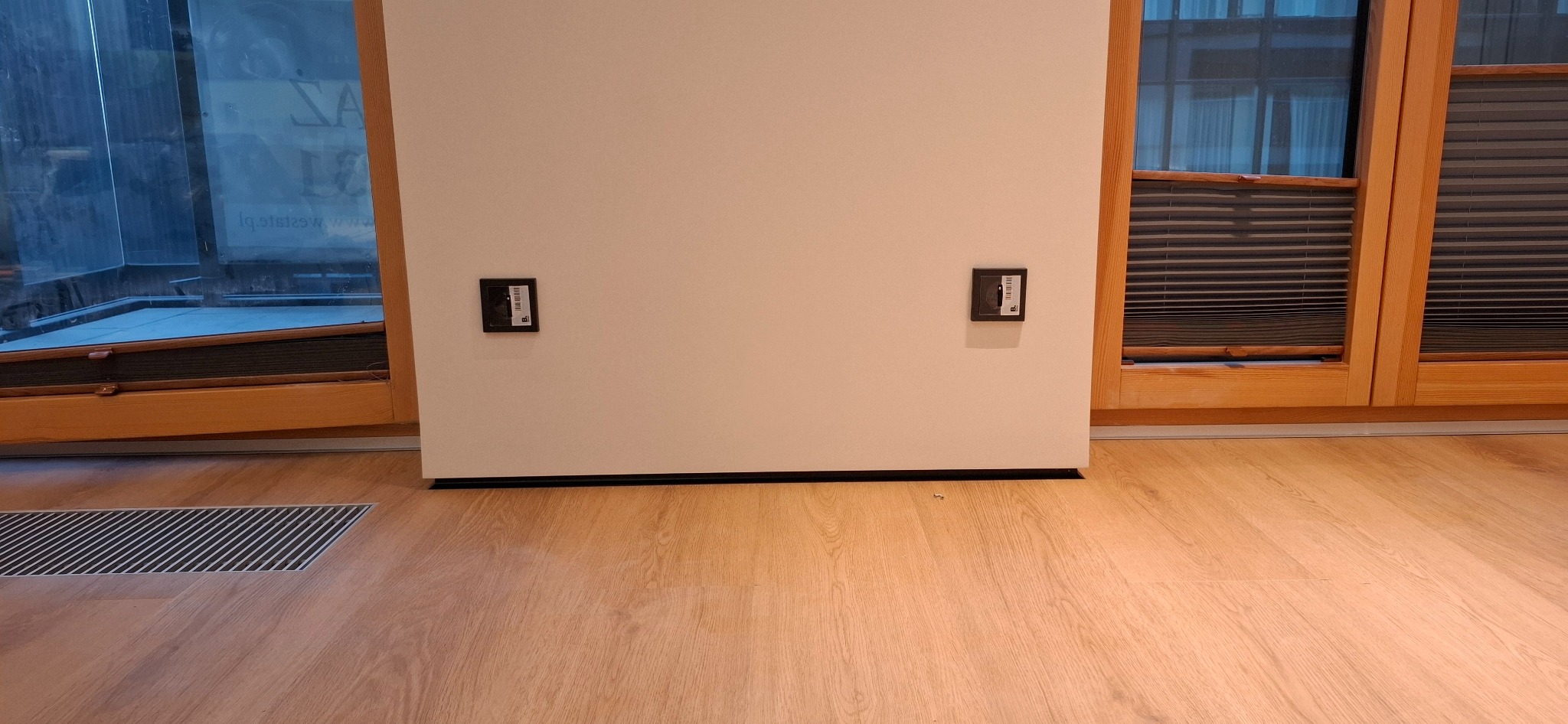 Jasna ściana z dwoma gniazdkami elektrycznymi, drewniana podłoga i okna z żaluzjami w tle. Widoczna kratka wentylacyjna w podłodze. Minimalistyczny design.