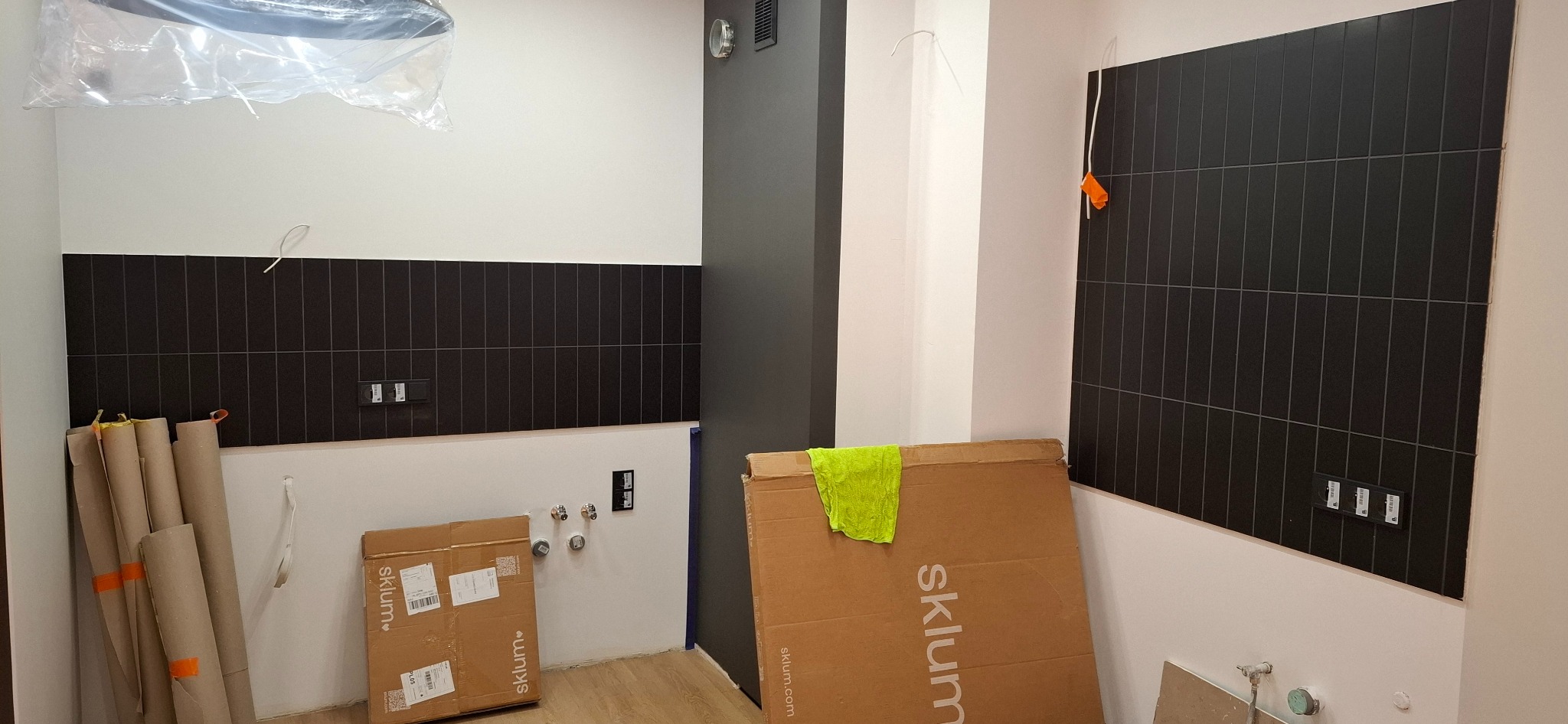 Częściowo wykończona ściana z czarnych, pionowych płytek w łazience. Widoczne kartony i rury, sugerujące trwający proces wykończeniowy.