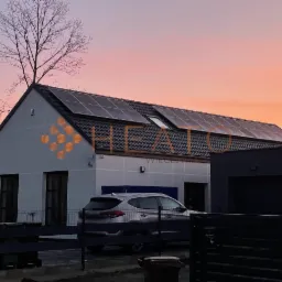 Nowoczesny dom jednorodzinny z panelami słonecznymi na dachu, widoczny samochód osobowy na podjeździe, brama wjazdowa i drzewo bez liści na tle zachodzącego słońca.