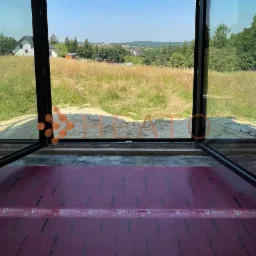 Wnętrze w trakcie budowy z otwartymi, dużymi oknami z widokiem na łąkę i zabudowania w oddali, podłoga pokryta czerwoną folią izolacyjną z widocznym napisem 'Red Snake'.