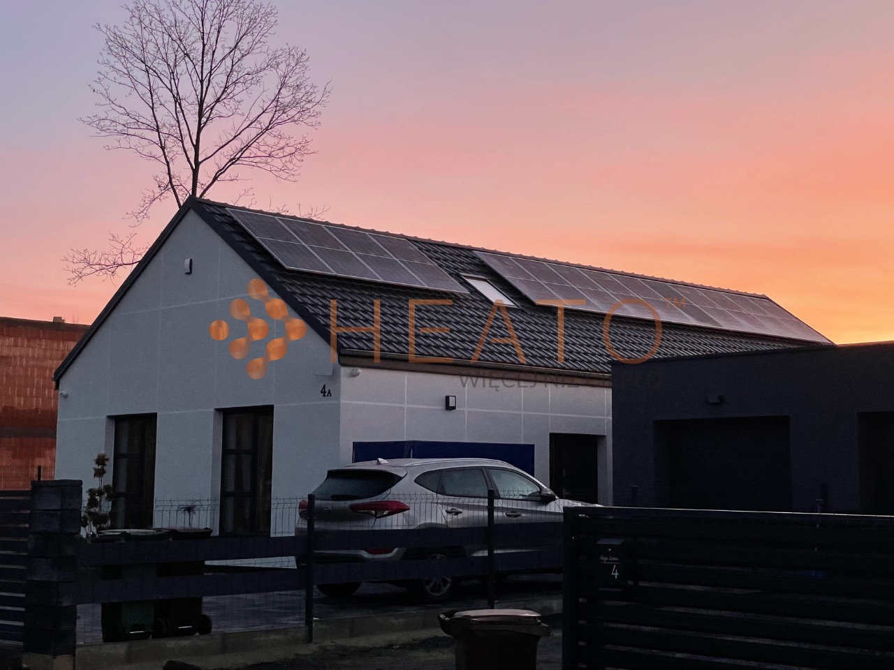 Nowoczesny dom jednorodzinny z panelami słonecznymi na dachu, widoczny samochód osobowy na podjeździe, brama wjazdowa i drzewo bez liści na tle zachodzącego słońca.