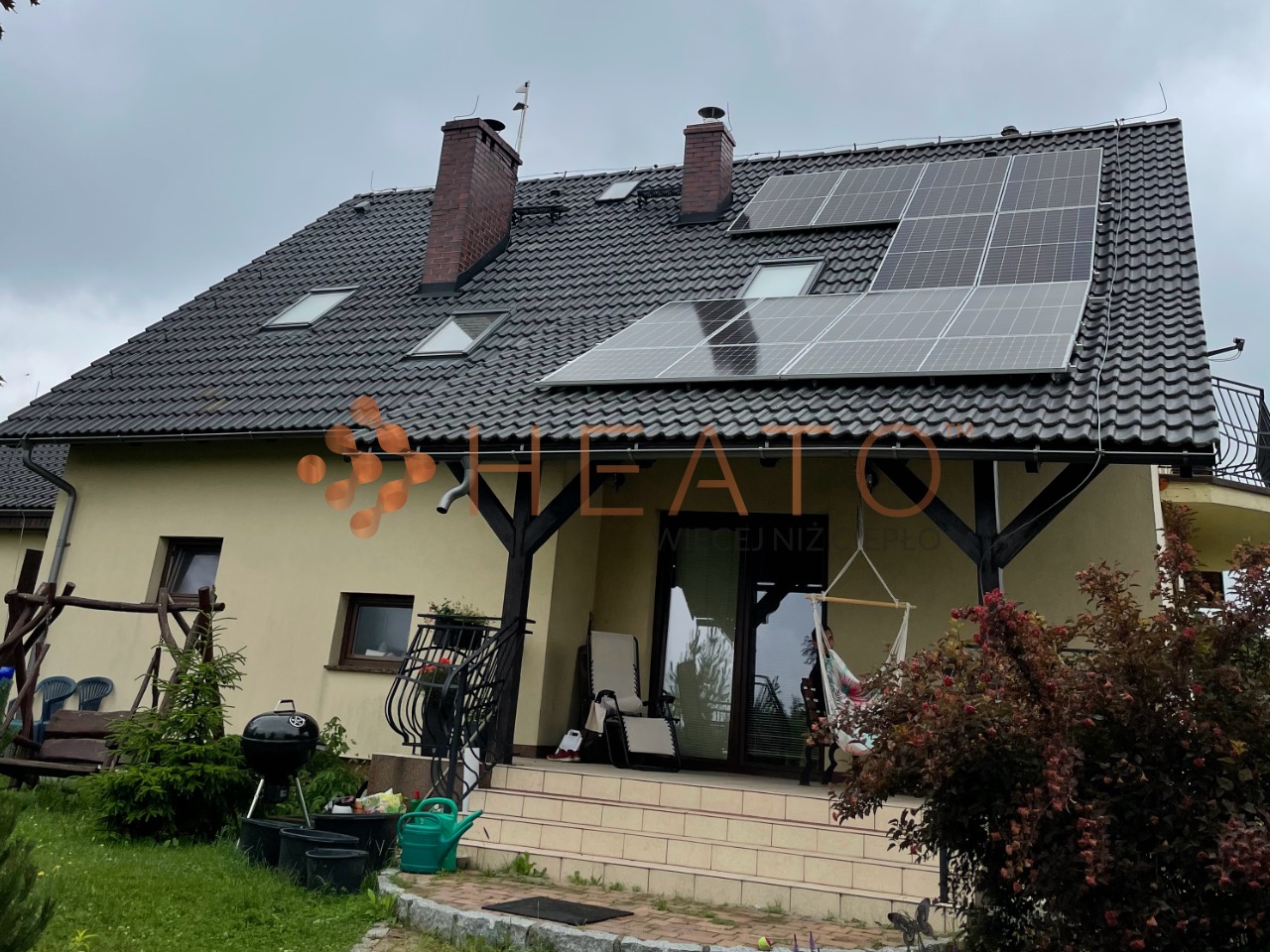 Dom jednorodzinny z panelami słonecznymi na dachu z dachówki ceramicznej, widoczne okna dachowe i komin, zadaszony taras z elementami drewnianymi, ogród z grillem i huśtawką.