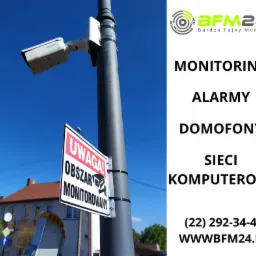 Kamera monitoringu zamontowana na słupie, widoczna tabliczka ostrzegawcza 'Uwaga! Obszar Monitorowany', w tle budynek z czerwonym dachem i błękitne niebo.