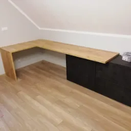 Drewniany blat roboczy w kształcie litery L, zamontowany w pokoju na poddaszu, wsparty na boku na drewnianej nodze, a z drugiej strony na czarnej komodzie z szufladami. Na komodzie leżą narzędzia...