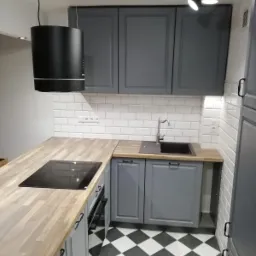 Szara zabudowa kuchenna z drewnianym blatem, czarną płytą indukcyjną i okrągłym okapem na tle białych płytek i czarno-białej podłogi w szachownicę.