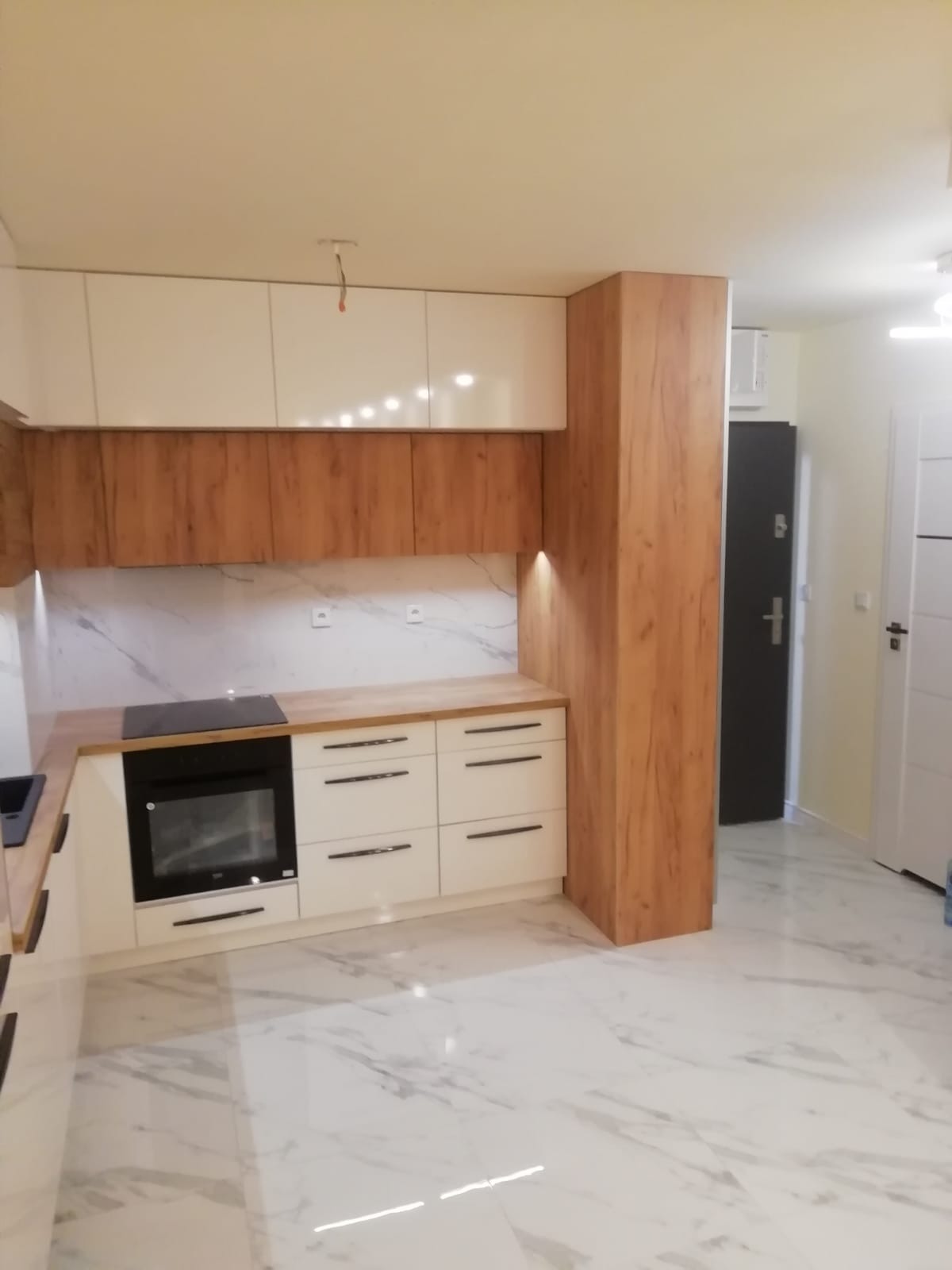 Nowoczesna kuchnia z białymi lakierowanymi szafkami górnymi i drewnopodobnymi szafkami dolnymi, blatem i obudową, z płytą indukcyjną i piekarnikiem w zabudowie, marmurowa ściana i podłoga.
