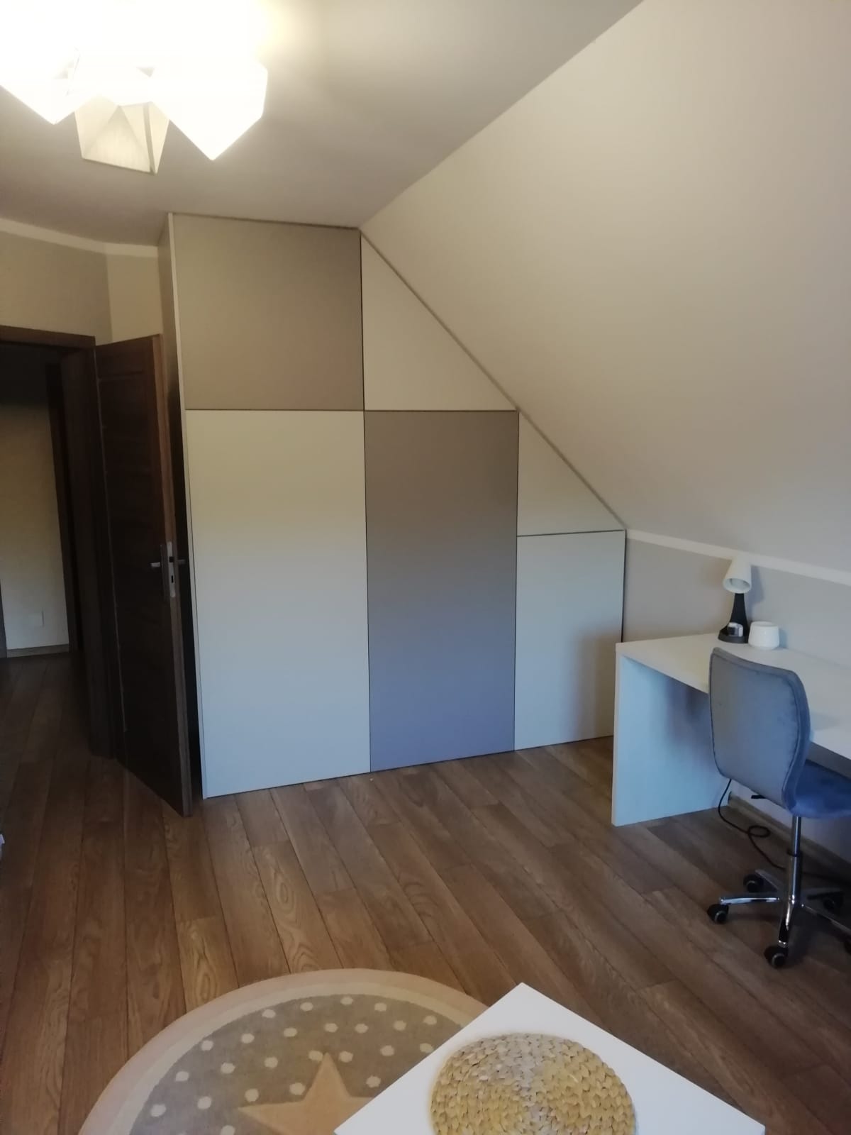 Pokój na poddaszu z zabudową szafy wnękowej z frontami w odcieniach szarości i bieli, biurko z krzesłem i lampką.