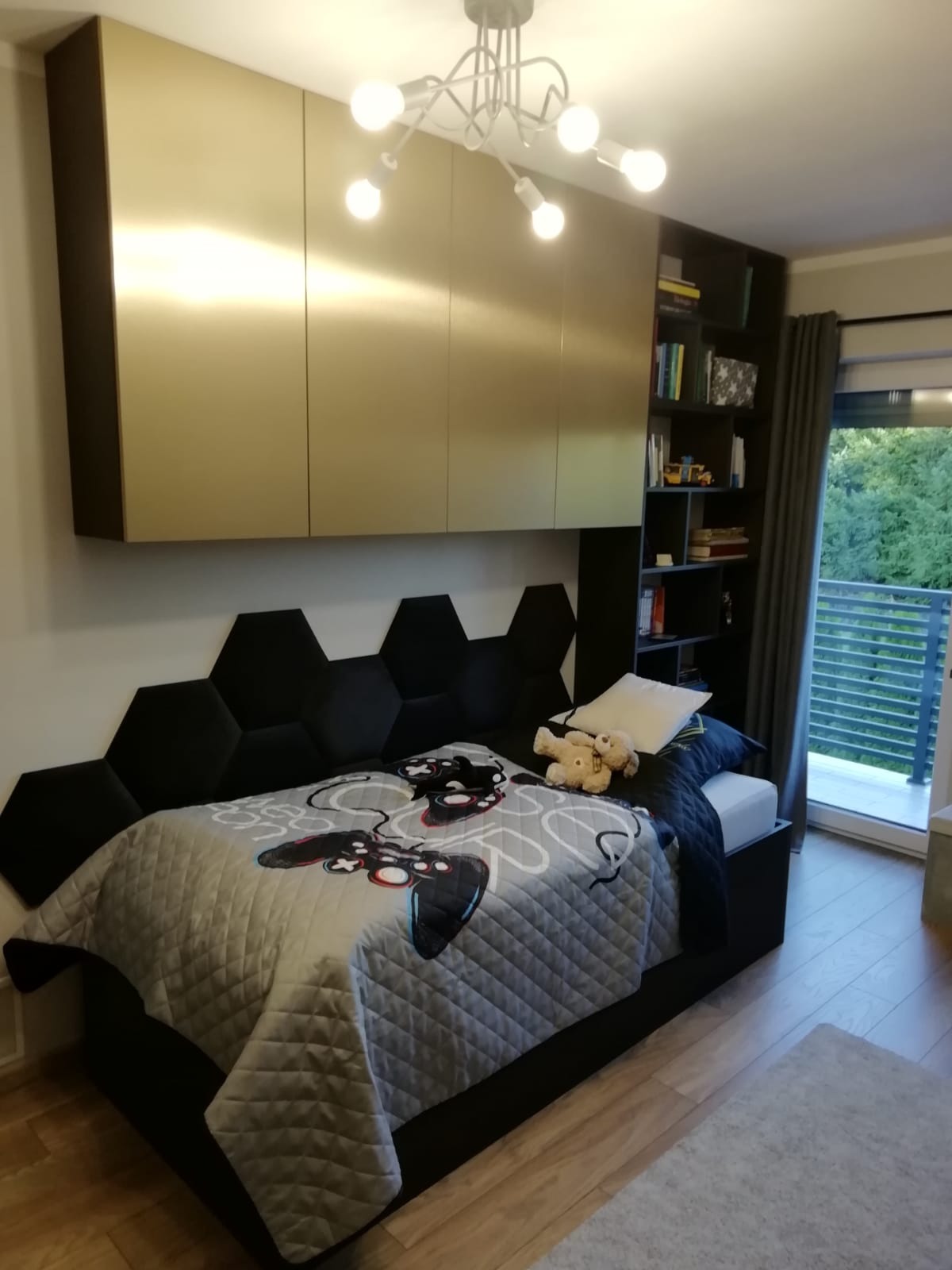 Pokój dziecięcy z łóżkiem, heksagonalnym zagłówkiem, szafkami wiszącymi w kolorze złotym i regałem na książki, oświetlony nowoczesną lampą sufitową.