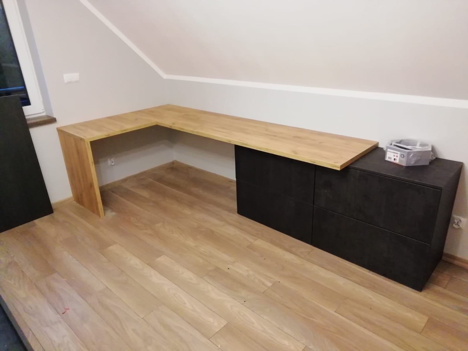 Drewniany blat roboczy w kształcie litery L, zamontowany w pokoju na poddaszu, wsparty na boku na drewnianej nodze, a z drugiej strony na czarnej komodzie z szufladami. Na komodzie leżą narzędzia...