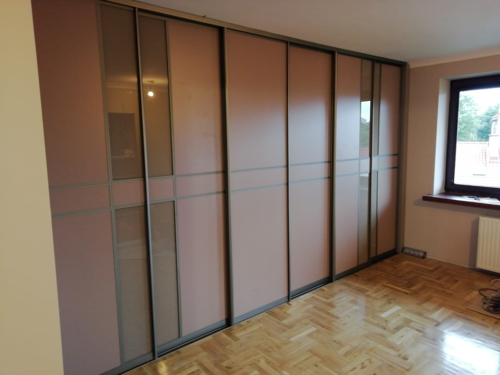 Wielodrzwiowa szafa wnękowa z przesuwnymi drzwiami w odcieniach różu i szarości, z podziałem na prostokątne panele i widocznym parkietem ułożonym w jodełkę.