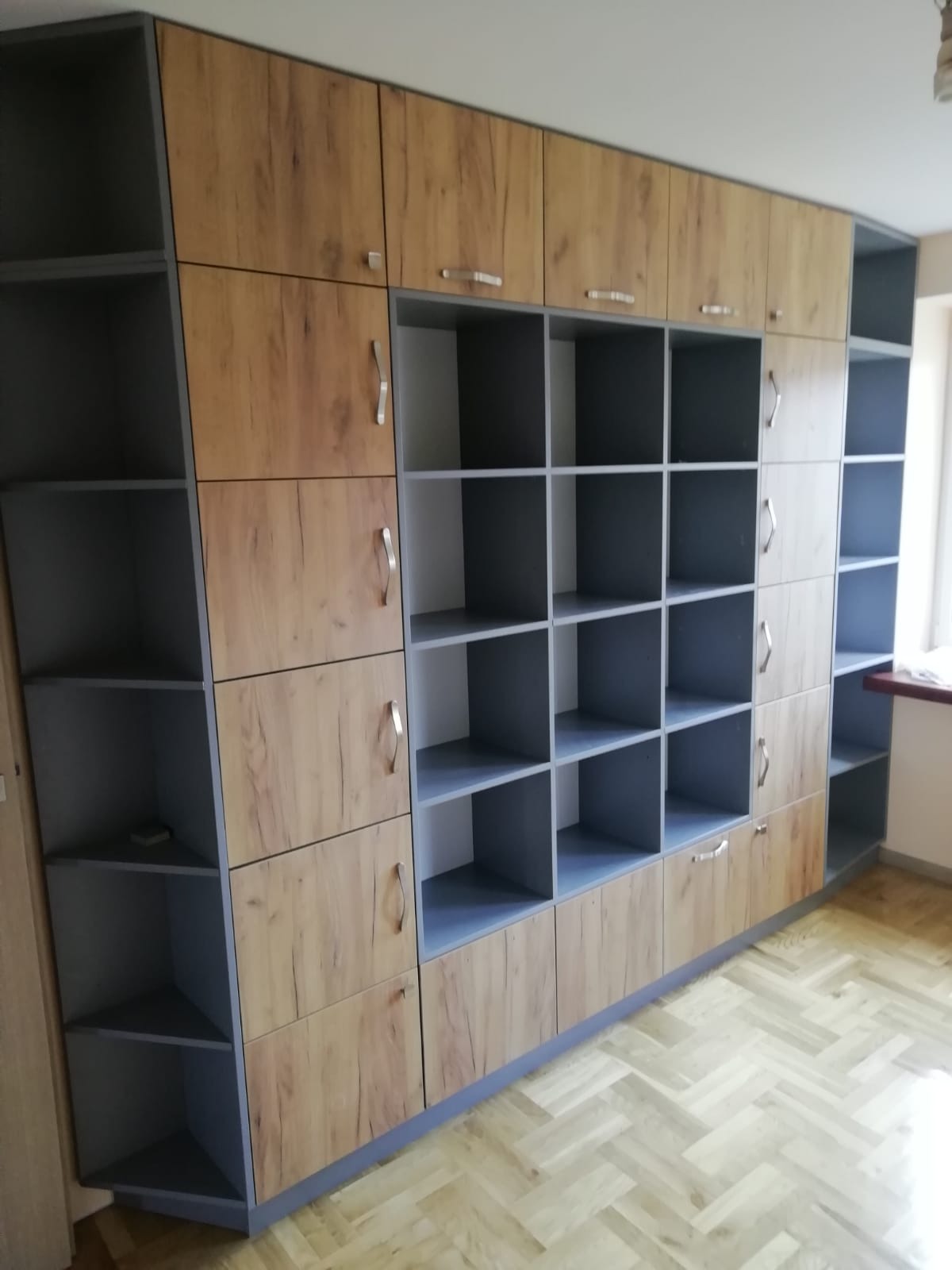 Zabudowa meblowa z szarymi półkami i frontami w kolorze drewna z widocznym usłojeniem, srebrne uchwyty, podłoga w jodełkę.