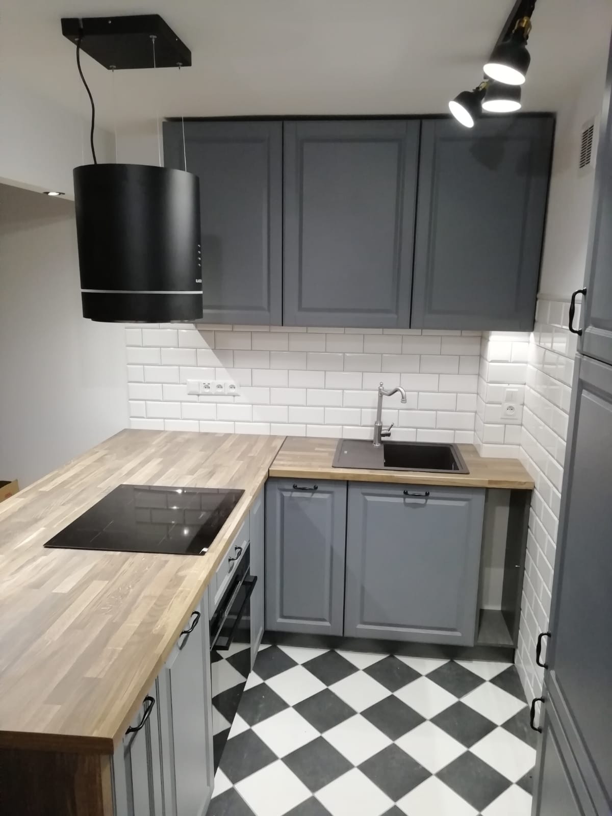 Szara zabudowa kuchenna z drewnianym blatem, czarną płytą indukcyjną i okrągłym okapem na tle białych płytek i czarno-białej podłogi w szachownicę.