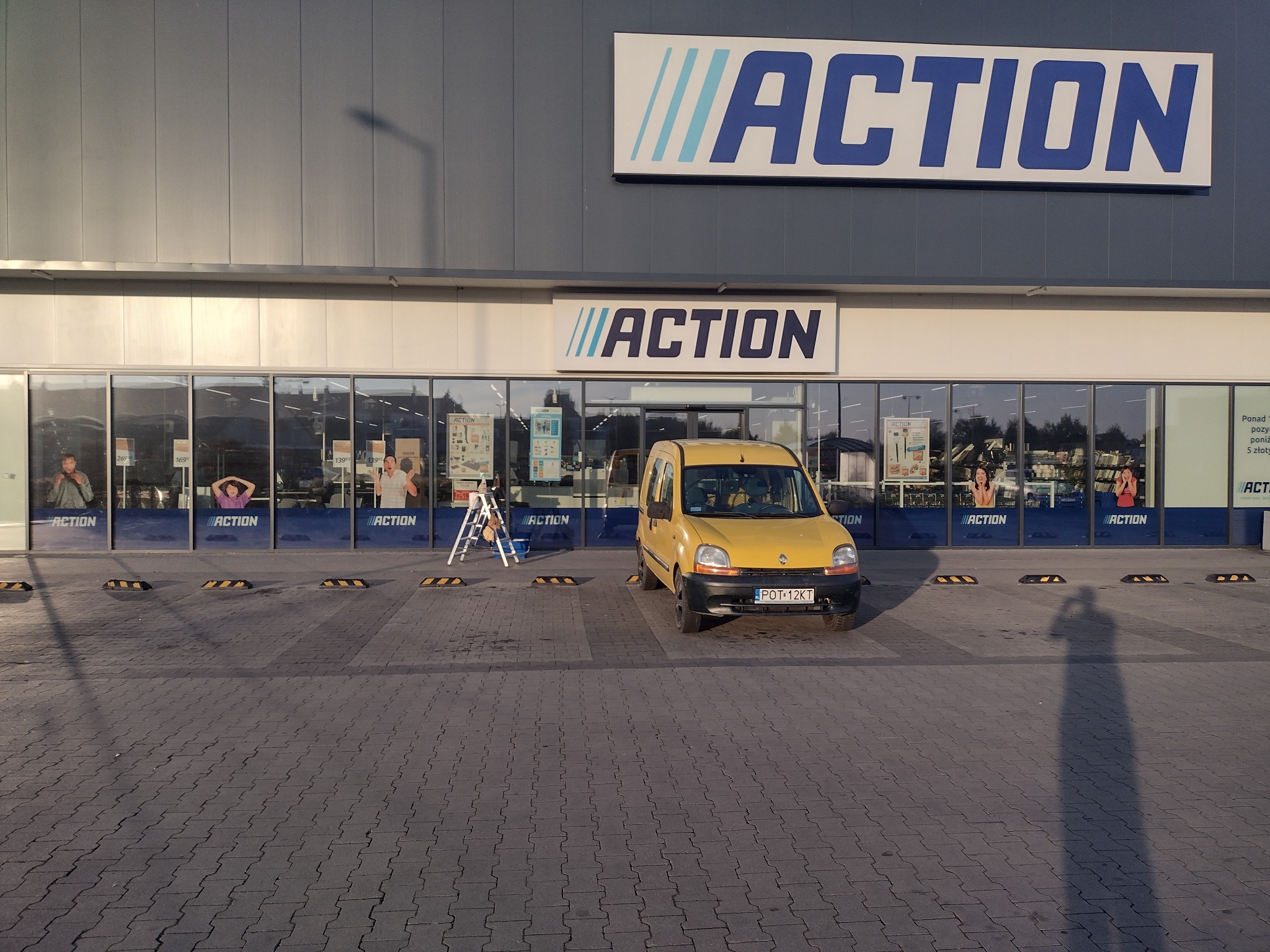 Żółty samochód dostawczy zaparkowany przed sklepem Action z drabiną opartą o witrynę, cień osoby robiącej zdjęcie widoczny na brukowanym parkingu.
