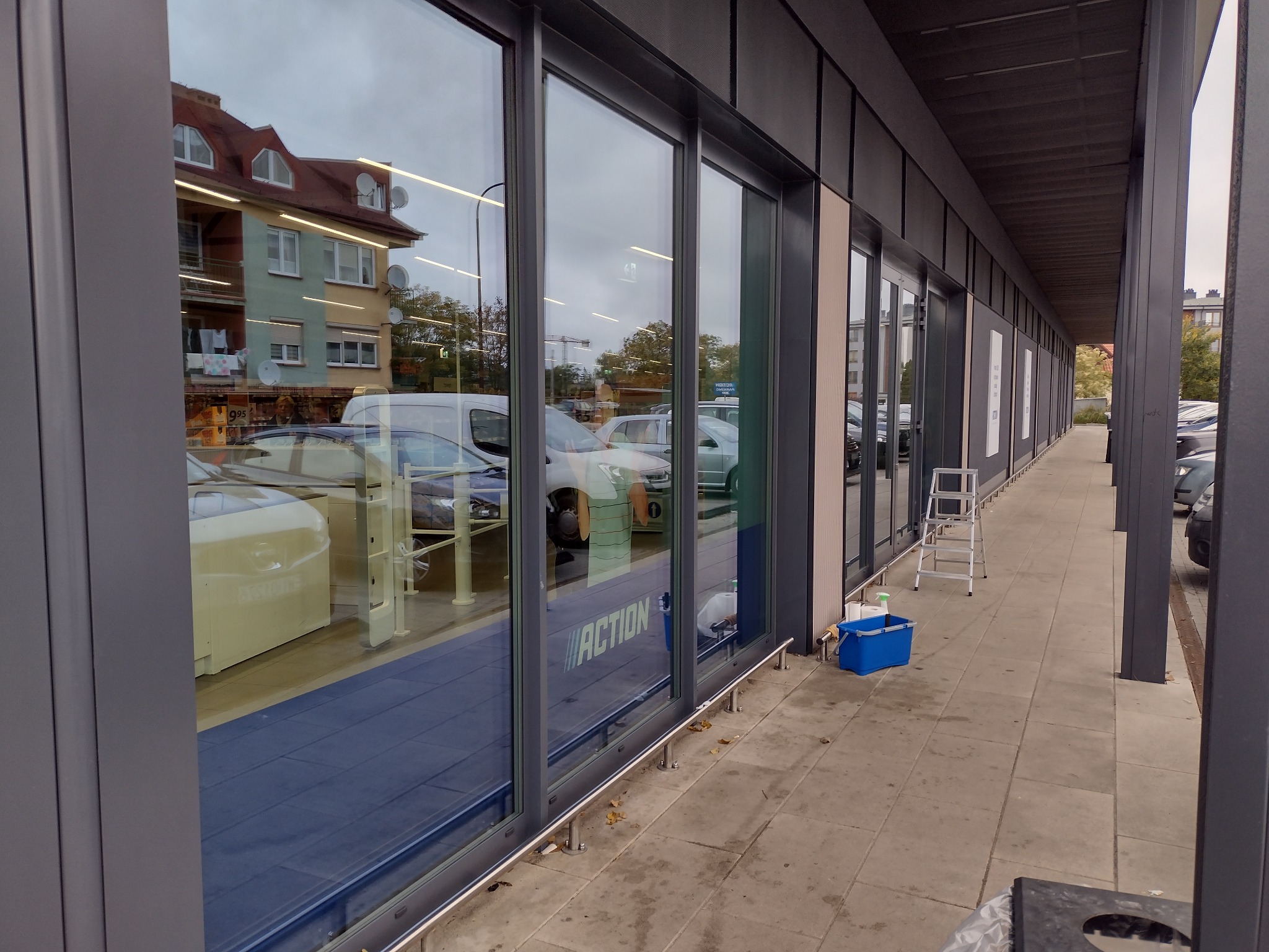 Zewnętrzne, szklane witryny sklepu z widocznym logo 'ACTION', częściowo umyte, z drabiną i niebieskim wiadrem z płynem do mycia okien obok.