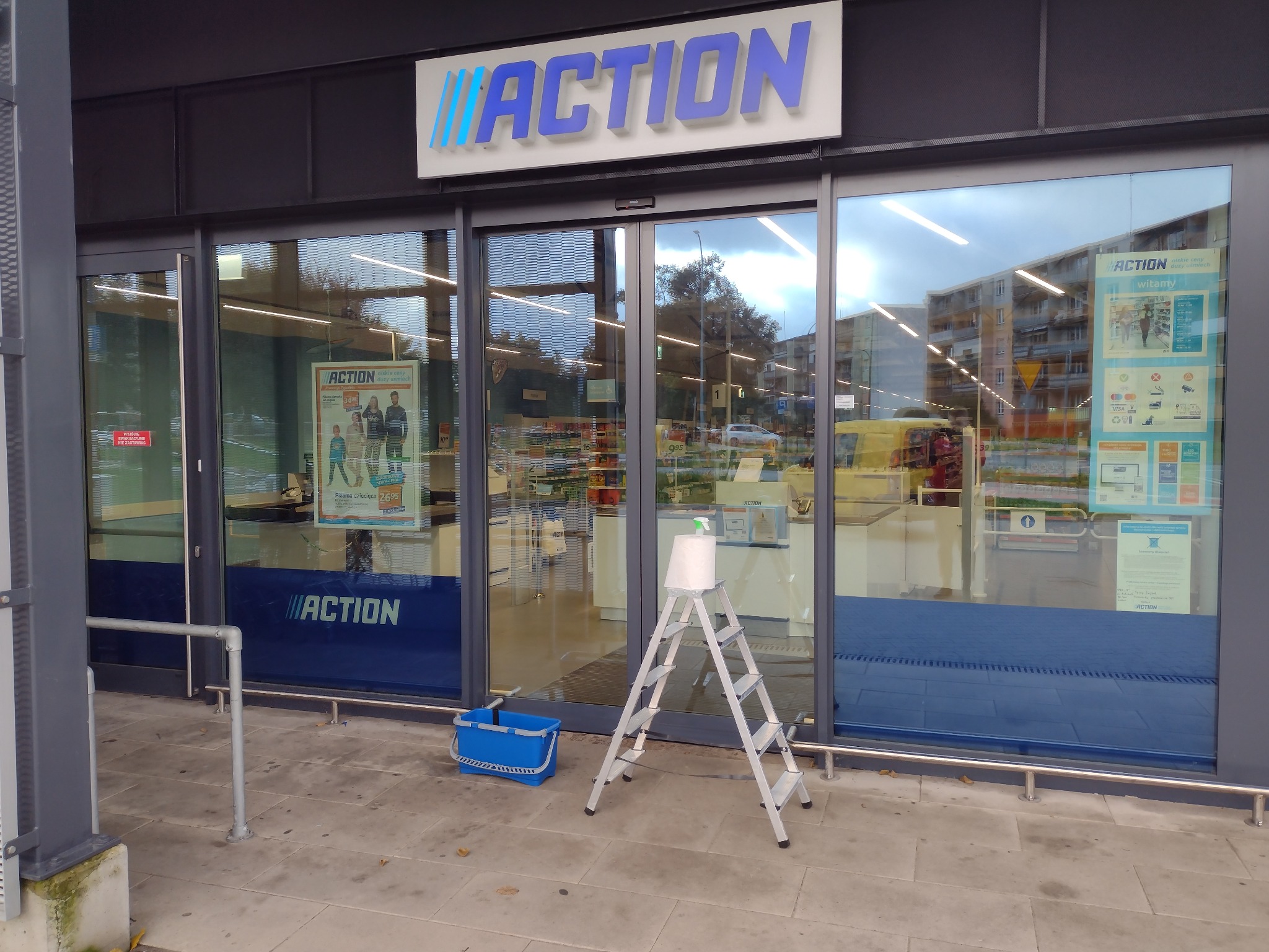 Aluminiowa drabina z rolką ręcznika papierowego i butelką ze spryskiwaczem na górnym stopniu, stojąca przed przeszklonym wejściem do sklepu Action, obok niebieski kubeł.
