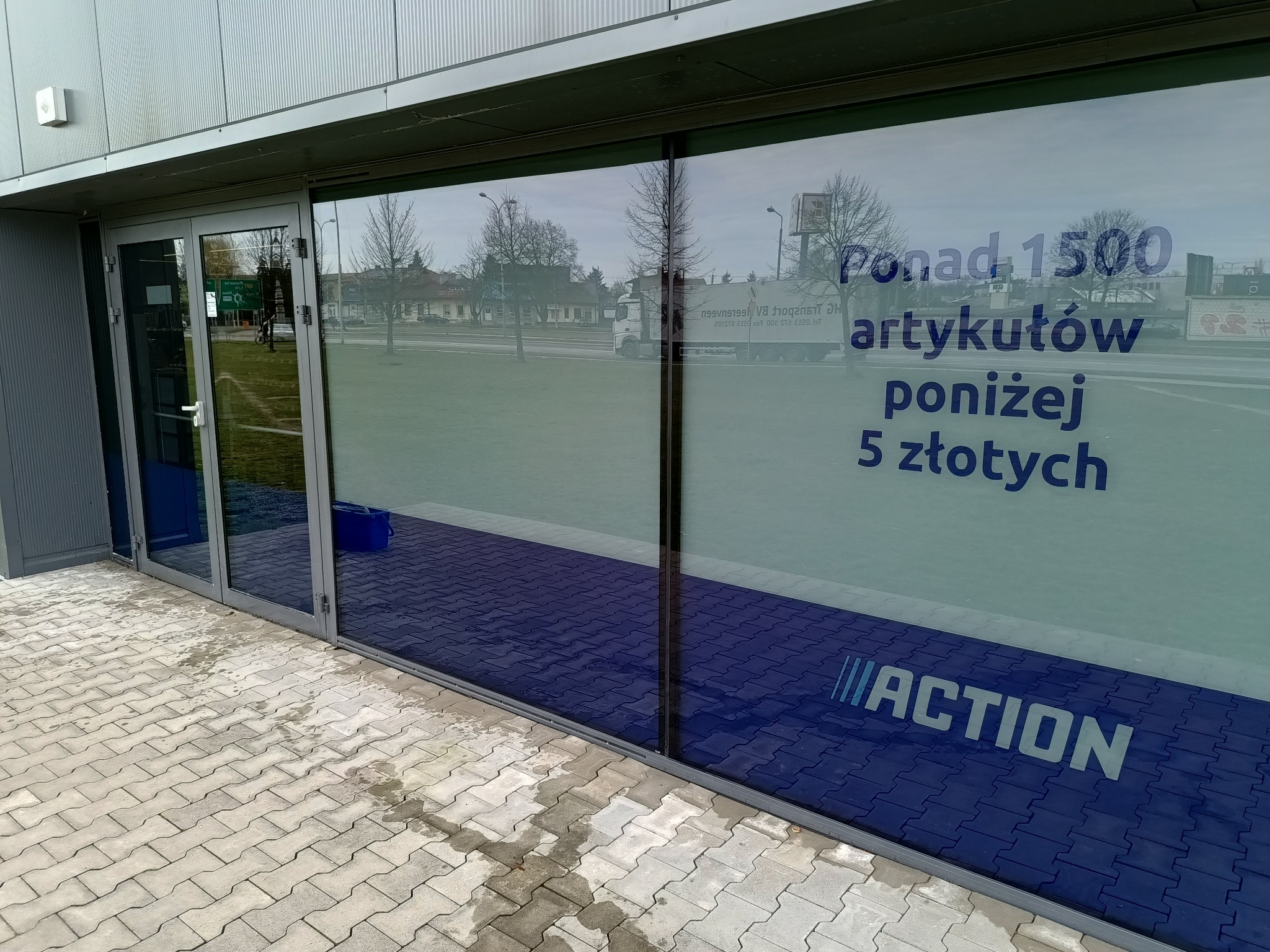 Szklana fasada sklepu z naklejonymi informacjami o promocji: 'Ponad 1500 artykułów poniżej 5 złotych' oraz logo 'ACTION', widoczne odbicie ulicy i drzew.