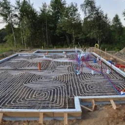 Fundament domu z rozłożonym systemem ogrzewania podłogowego, widoczne białe rurki ułożone w spiralny wzór wewnątrz drewnianego szalunku, z wystającymi rurkami instalacji wodnej i kanalizacyjnej...