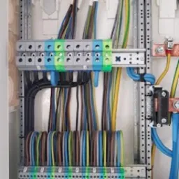 Rozdzielnica elektryczna z precyzyjnie ułożonymi przewodami o różnych kolorach, podłączonymi do listew zaciskowych. Widoczne metalowe szyny montażowe i elementy mocujące. Przewody ułożone...