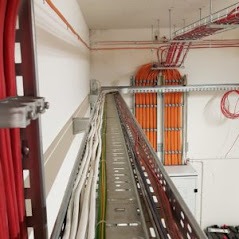 Koryto kablowe wypełnione przewodami elektrycznymi w instalacji przemysłowej, widok z perspektywy, przewody w kolorach czerwonym, pomarańczowym, zielonym i żółtym, metalowe wsporniki mocujące...