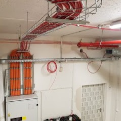 Koryto kablowe z pomarańczowymi przewodami ułożonymi w równoległe rzędy, zamontowane pod sufitem w pomieszczeniu technicznym. Dodatkowe przewody prowadzone po ścianach, widoczna szafa sterownicza...