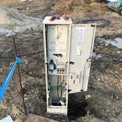 Otwarta skrzynka elektryczna z narzędziami na górze, umieszczona w wykopie ziemnym. Widoczne kable i elementy instalacji wewnątrz skrzynki. Obok skrzynki wbity pręt z niebieską taśmą.